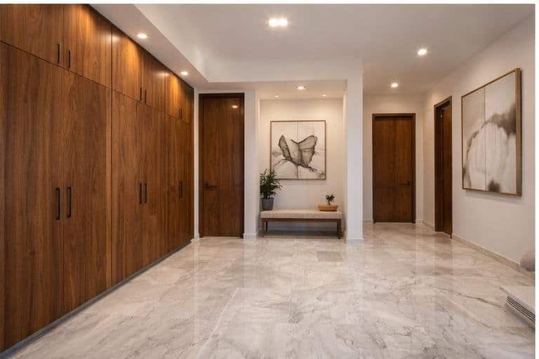 Pasillo interior con pisos de mármol, puertas y closets de madera sólida en acabado natural, iluminación indirecta y estilo moderno elegante.